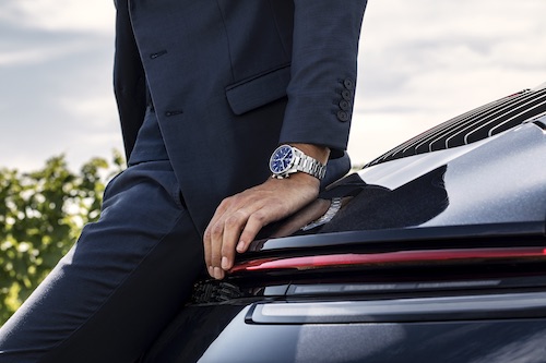 Image of a man in a blue suit wearing a TAG-Heuer Carrera watch while leaning against a Porsche Carrerra sports car. 